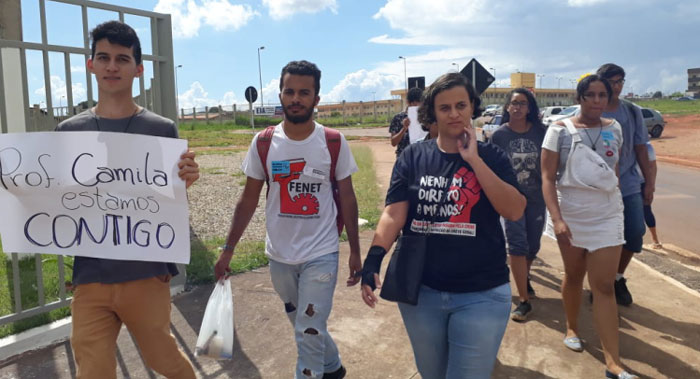 Foto da professora Camila Marques com seus alunos em volta. Um deles carrega um cartaz escrito "Prof Camila estamos contigo"