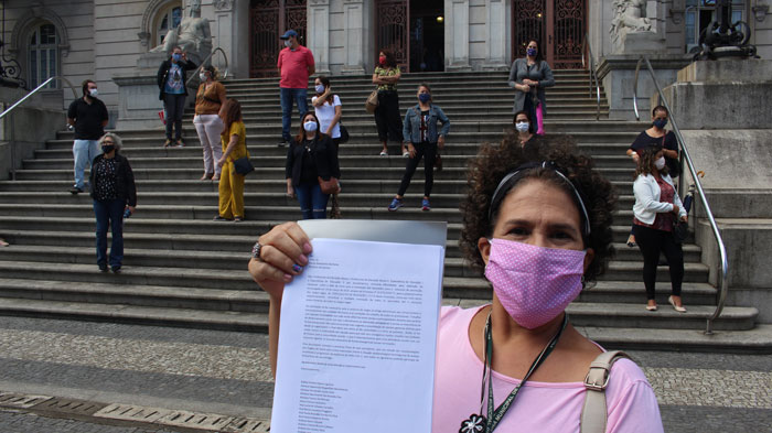Foto com uma servidora mostrando a carta em primeiro plano e no fundo outras servidoras na escadaria do Paço Municipal respeitando o distanciamento