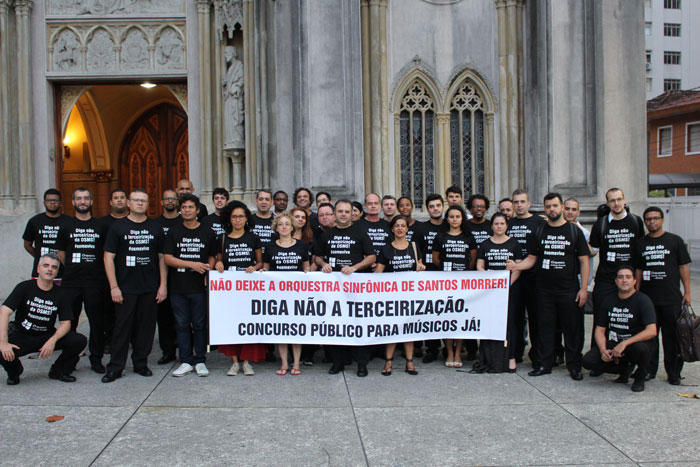 Músicos da orquestra em frente à igreja do Embaré com a faixa "NÃO DEIXE A ORQUESTRA SINFÔNICA DE SANTOS MORRER! DIGA NÃO A TERCEIRIZAÇÃO! CONCURSO PÚBLICO PARA MÚSICOS JÁ!"
